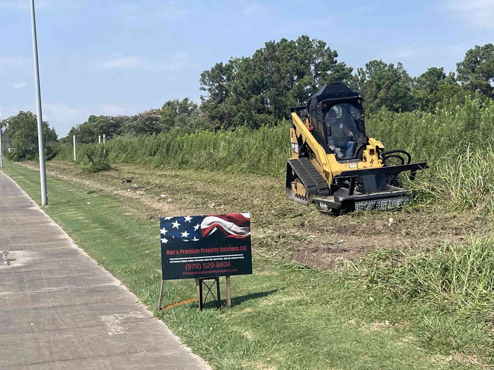 A tracked skid steer with a mulching head clearing brush beside a Ron's Premium Property Solutions job sign