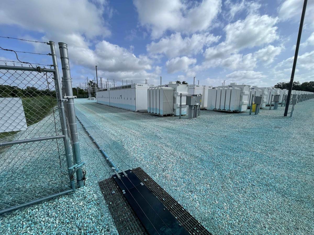 Battery storage containers and transformers behind a chain link gate on a graveled yard