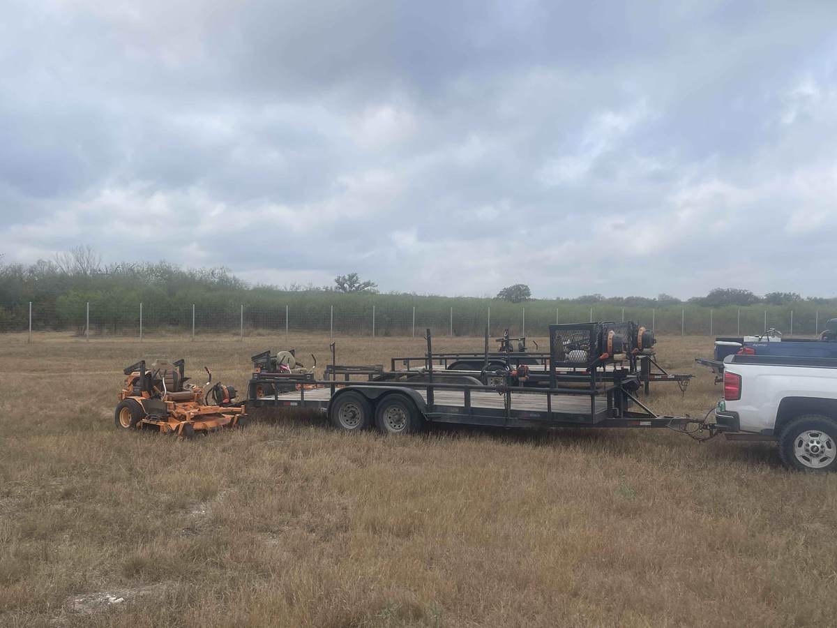 Zero-turn mowers, trimmers, and blowers on trailers behind a work truck at a fenced site