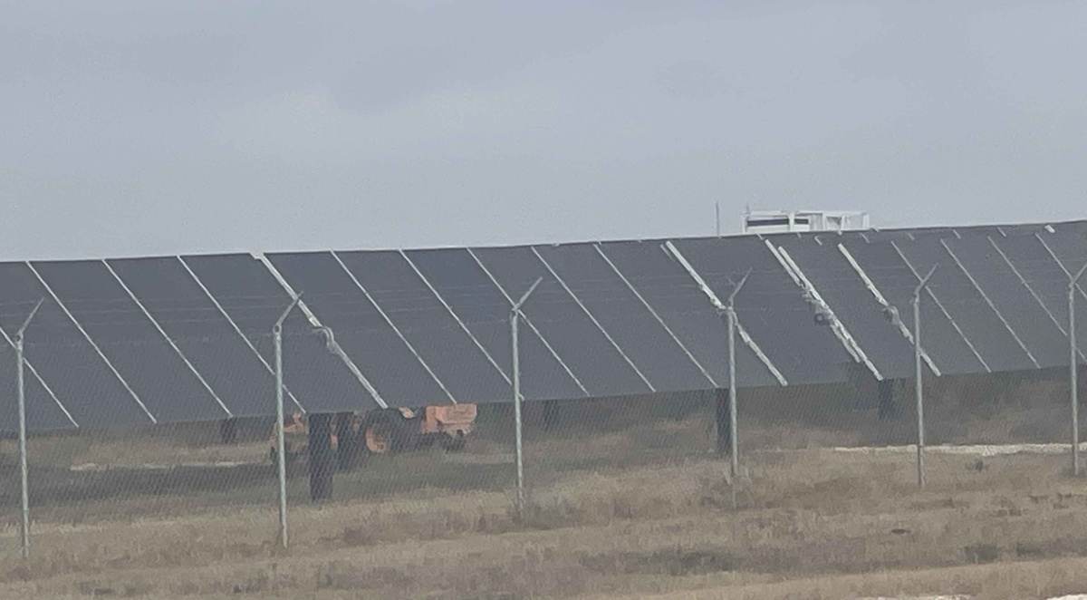 Rows of solar panels behind a perimeter fence with equipment working underneath